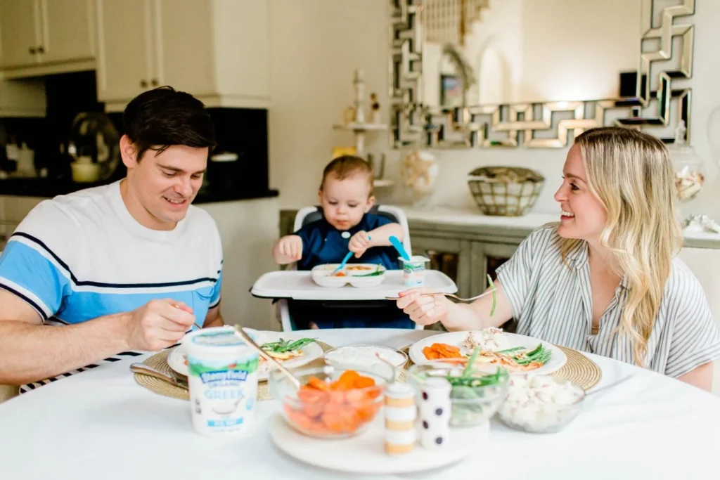 A couple shares a meal at a table, with a baby seated in a high chair beside them.