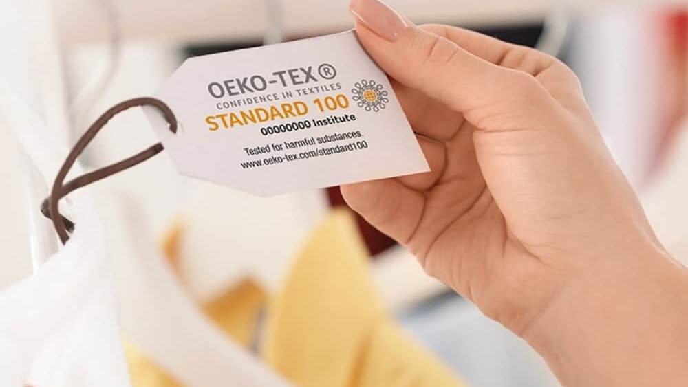 A woman holding an OEKO-TEX standard 100 tag while examining clothes on a rack in a retail store.