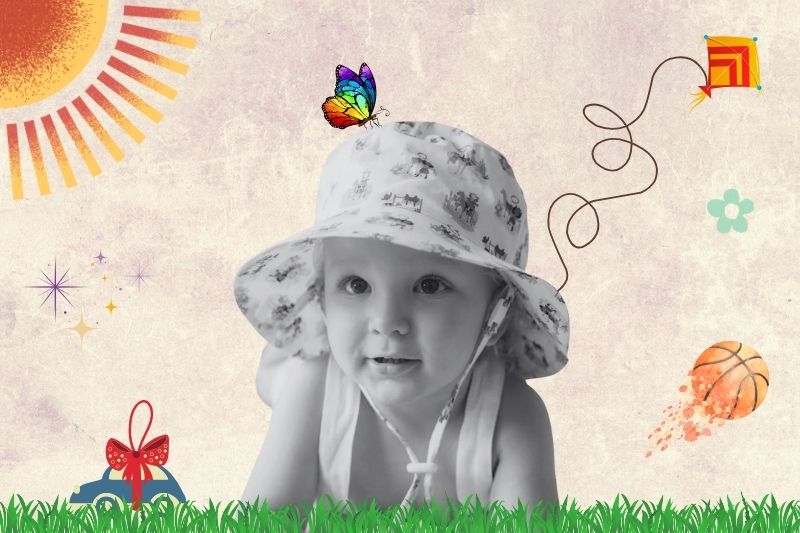 A baby wearing a hat sits on green grass, smiling and enjoying a sunny day outdoor.