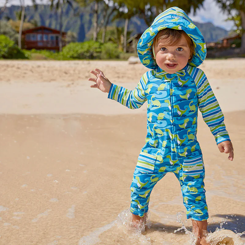 A baby boy is standing in water on a beach in a swimsuit.
