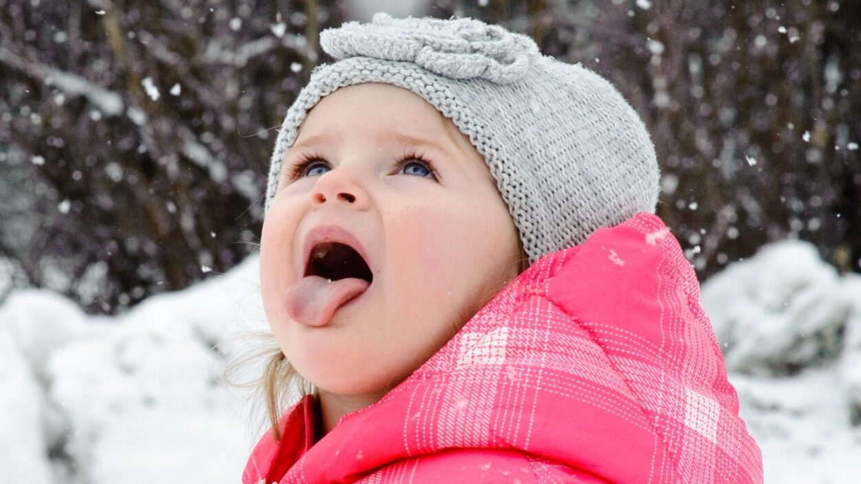 A young girl with her mouth open stands in the snow, expressing joy or amazement in a winter scene.