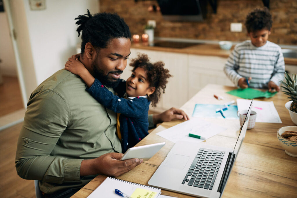A father and his two children are seated at a table, engaging with a laptop in a cozy indoor setting.