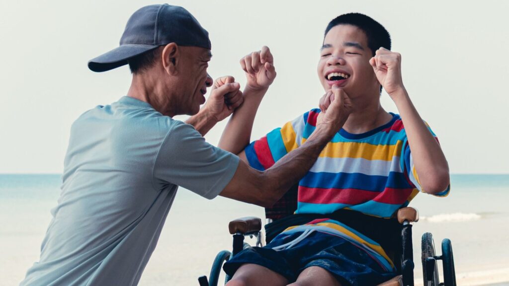 A man in a wheelchair assists a boy playing on the beach, fostering an inclusive and supportive environment.