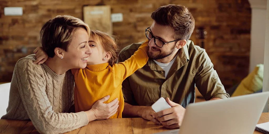 A family of three, a husband, wife, and child, gather around a table with a laptop, sharing a moment of love and compassion.