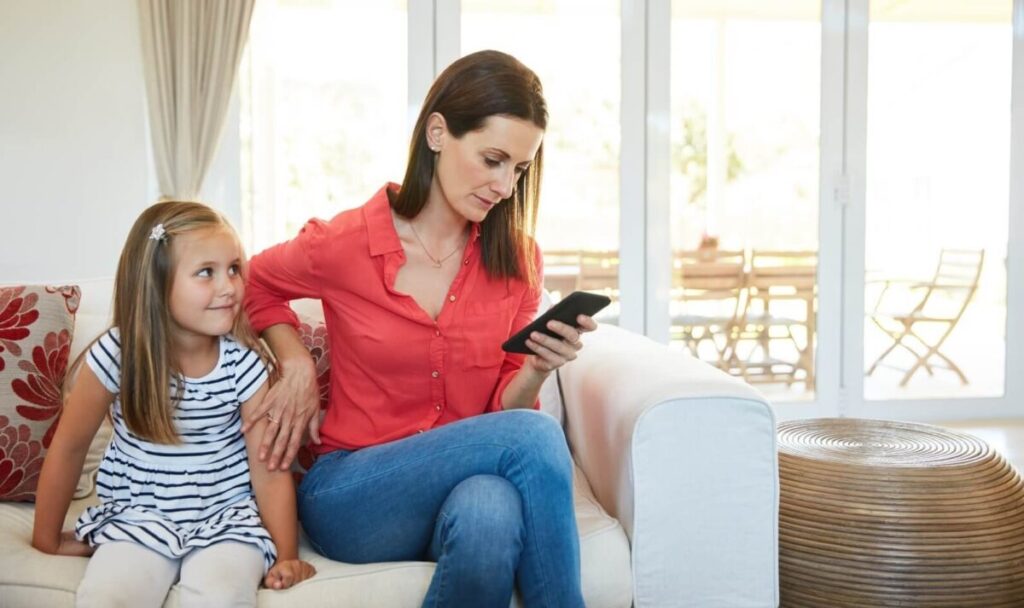 A mother and her daughter are seated on a couch. The mother is looking at her phone, ignoring her daughter.
